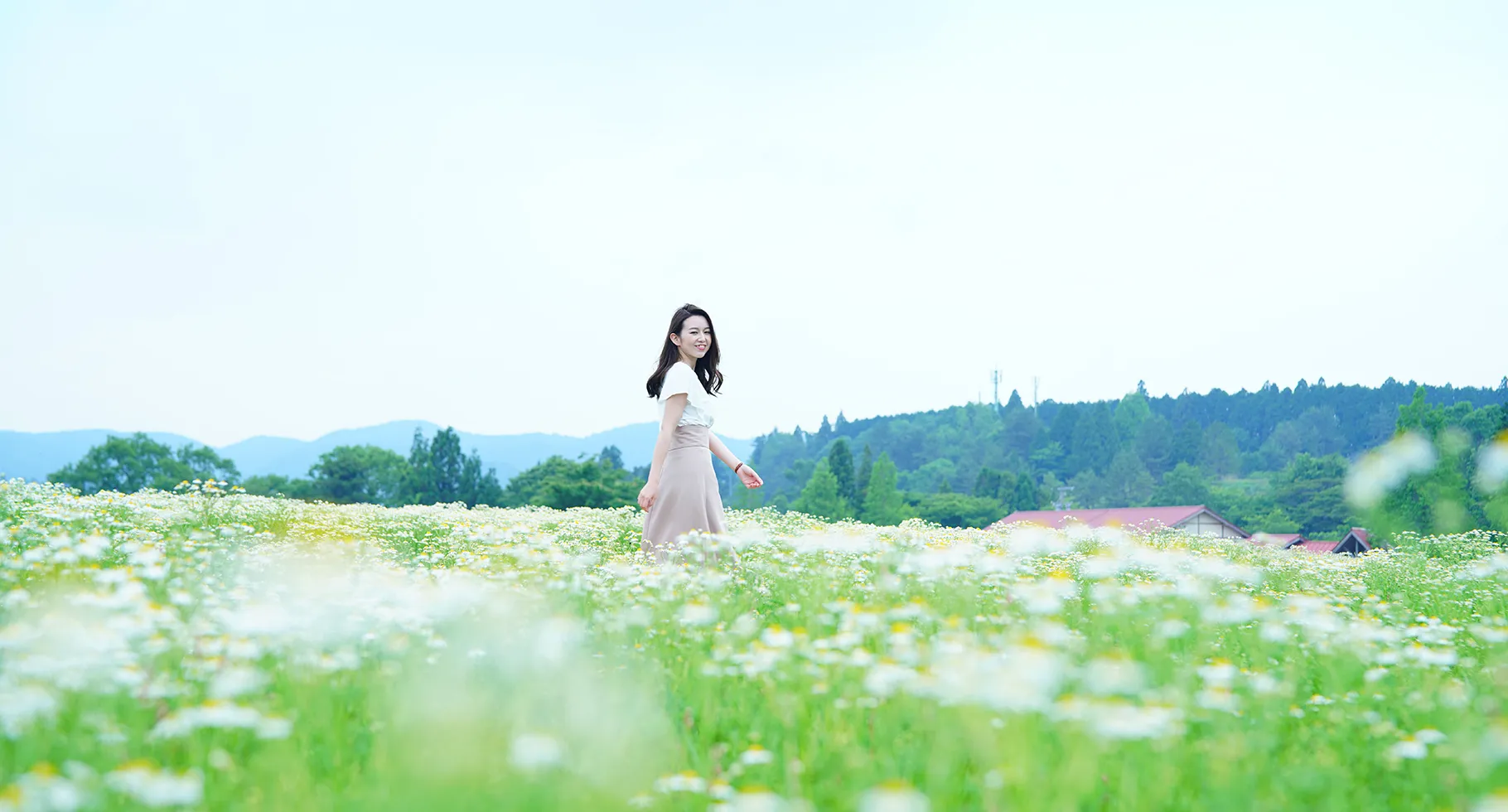 花畑の中を歩く女性。背景には緑豊かな山々と家が見える、自然に囲まれた風景。